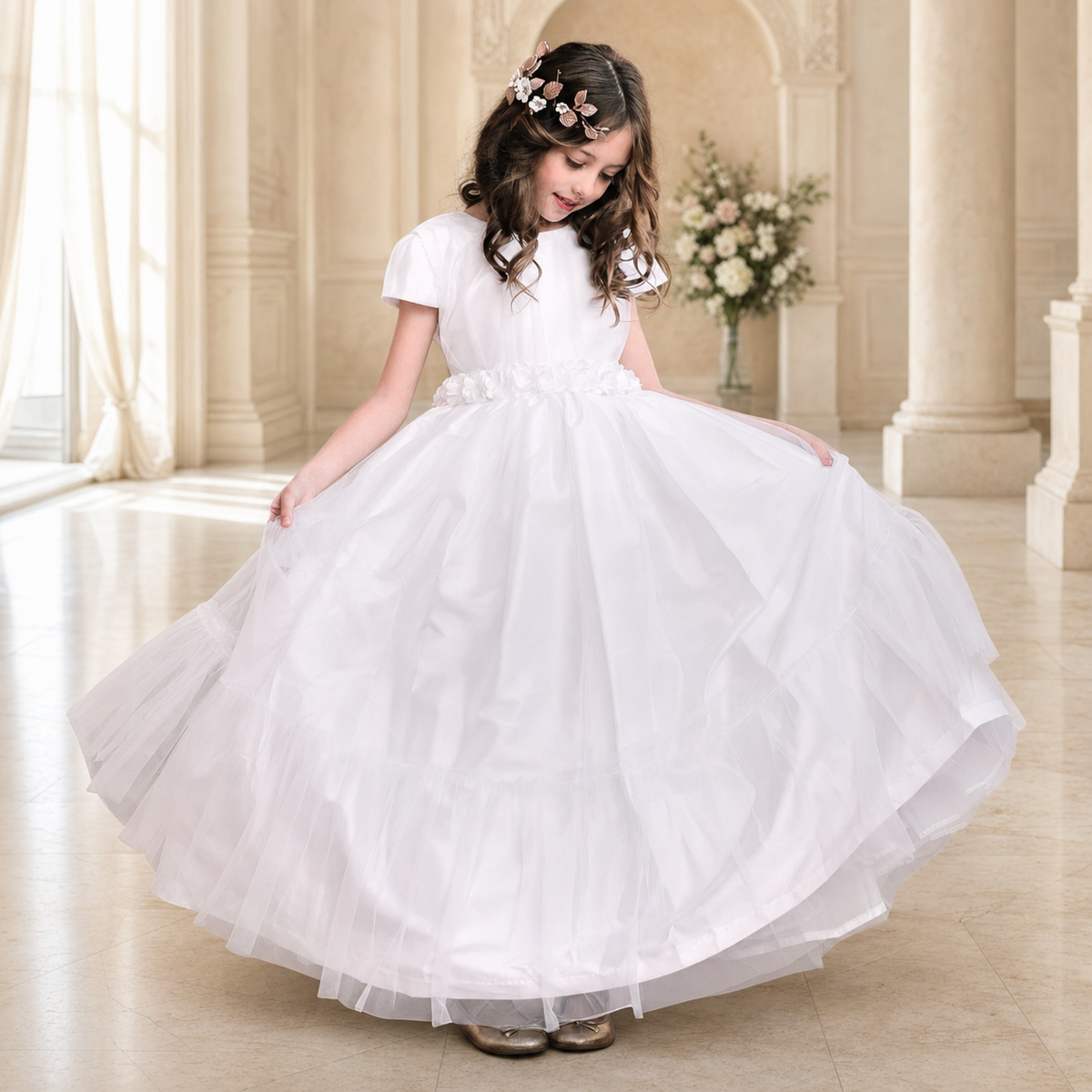 Girl wearing the white Clara First Communion dress with a floral waistband and layered tulle skirt in an elegant cream interior