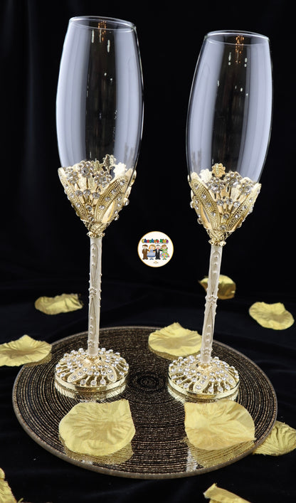 Two elegant Wedding champagne flutes with decorative bases on a black surface with gold leaves.