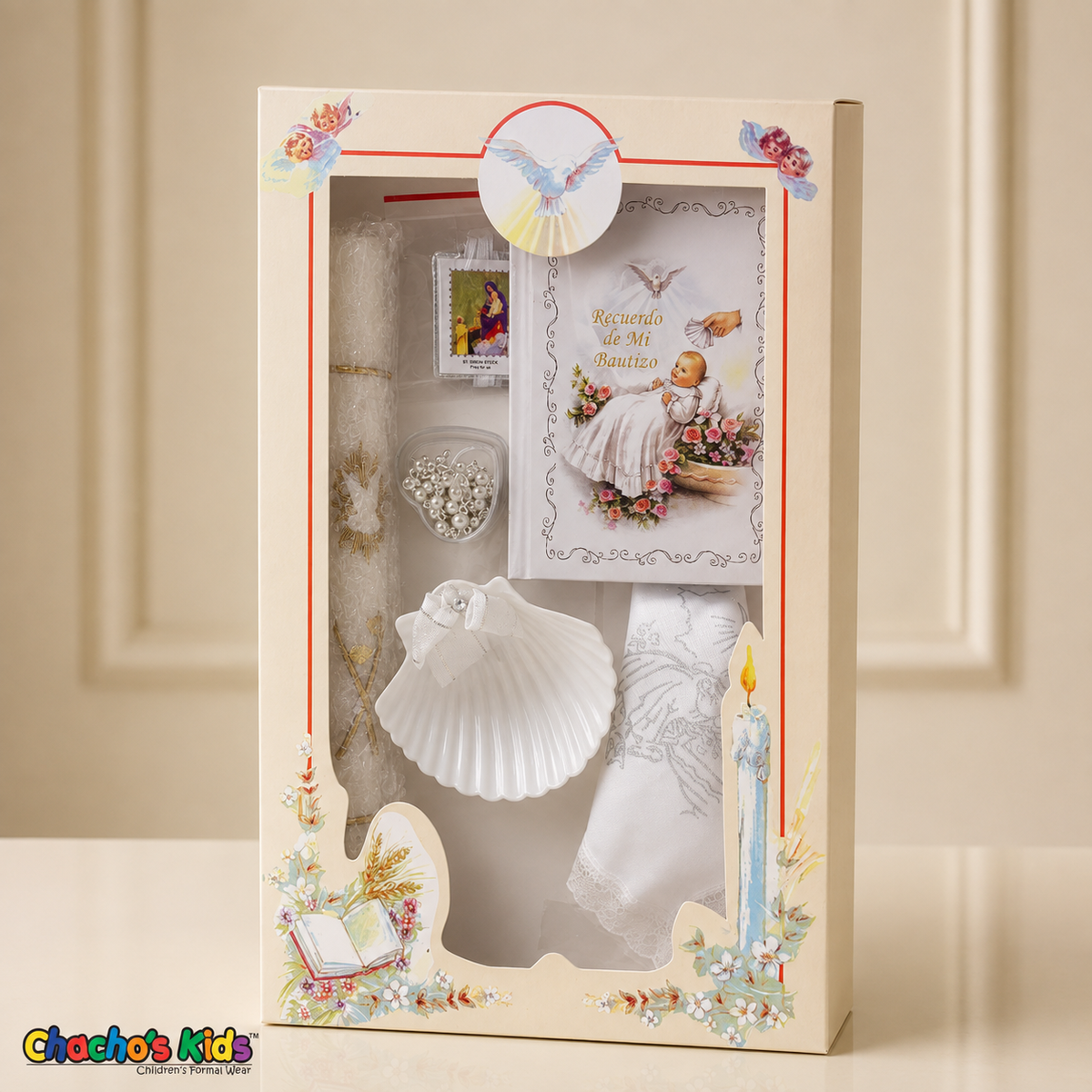 White six-piece Spanish baptism candle set standing in its illustrated window gift box on a cream tabletop, with prayer book, shell, rosary, scapular and handkerchief.
