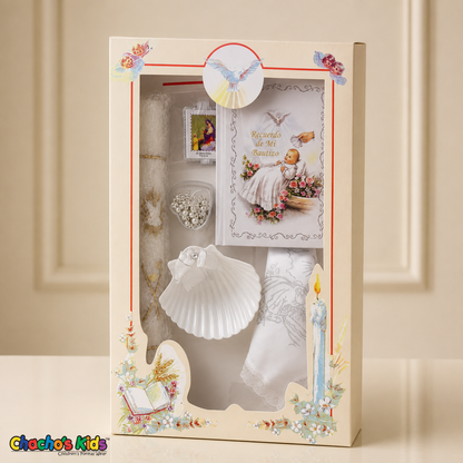 White six-piece Spanish baptism candle set standing in its illustrated window gift box on a cream tabletop, with prayer book, shell, rosary, scapular and handkerchief.