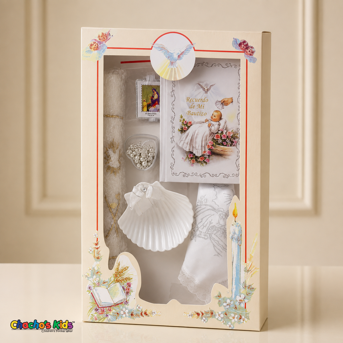 White six-piece Spanish baptism candle set standing in its illustrated window gift box on a cream tabletop, with prayer book, shell, rosary, scapular and handkerchief.