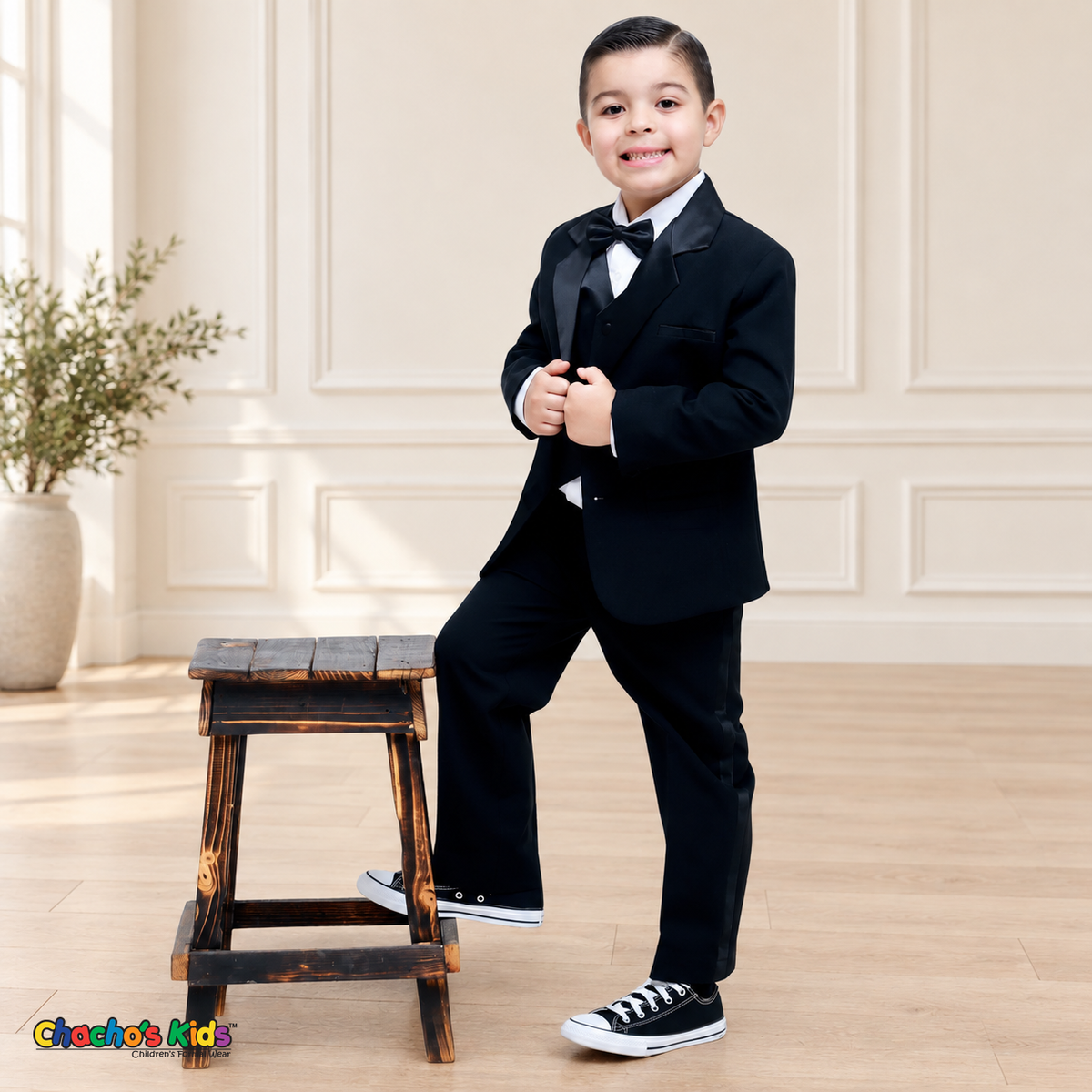 Boys Signature Tuxedo in Black (BY010), child model standing beside a wooden stool with satin lapels and a black bow tie.