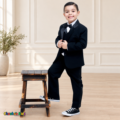 Boys Signature Tuxedo in Black (BY010), child model standing beside a wooden stool with satin lapels and a black bow tie.