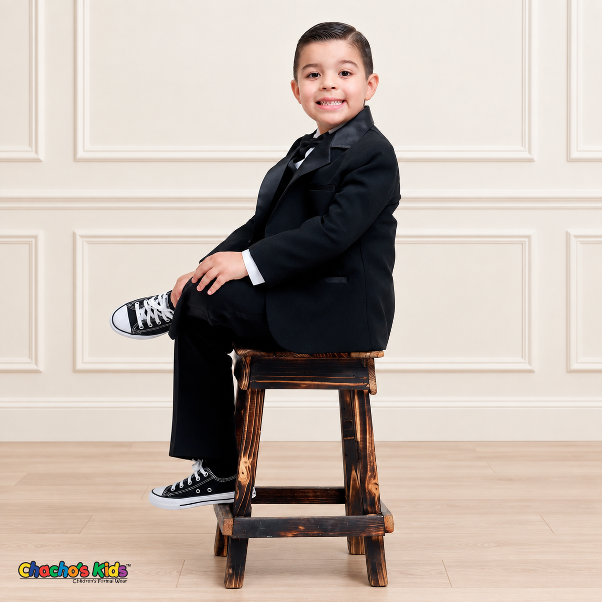 Boys Signature Tuxedo in Black (BY010), child model seated on a wooden stool with crossed legs, wearing a black tuxedo and white shirt.