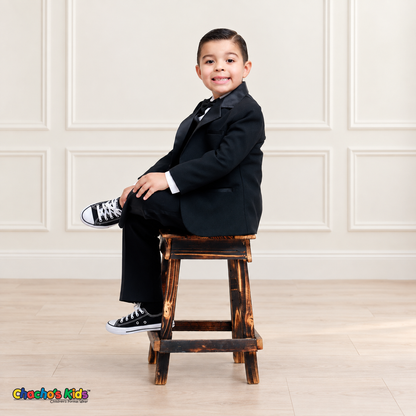 Boys Signature Tuxedo in Black (BY010), child model seated side pose on a wooden stool, showing the black tuxedo jacket and trousers.
