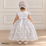 Baby girl wearing a white floral lace baptism dress and matching bonnet, back without cape, showing the ribbon bow and embroidered skirt