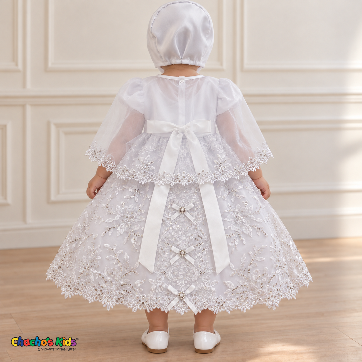 Baby girl wearing the white Virgin Mary Baptism Dress, Style 374, and matching satin bonnet, back view with the sheer embroidered cape, showing the satin sash, floral embroidery and ribbon bows.