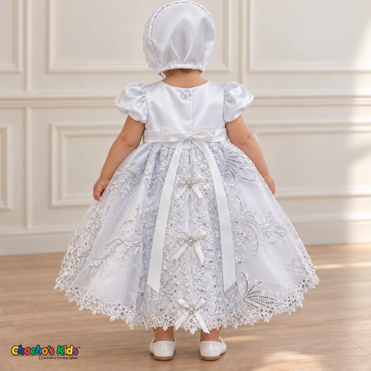 Baby girl wearing the white Virgin Mary Baptism Dress, Style 374, and matching satin bonnet, back view without the cape, showing the satin sash, floral embroidery and ribbon bows.