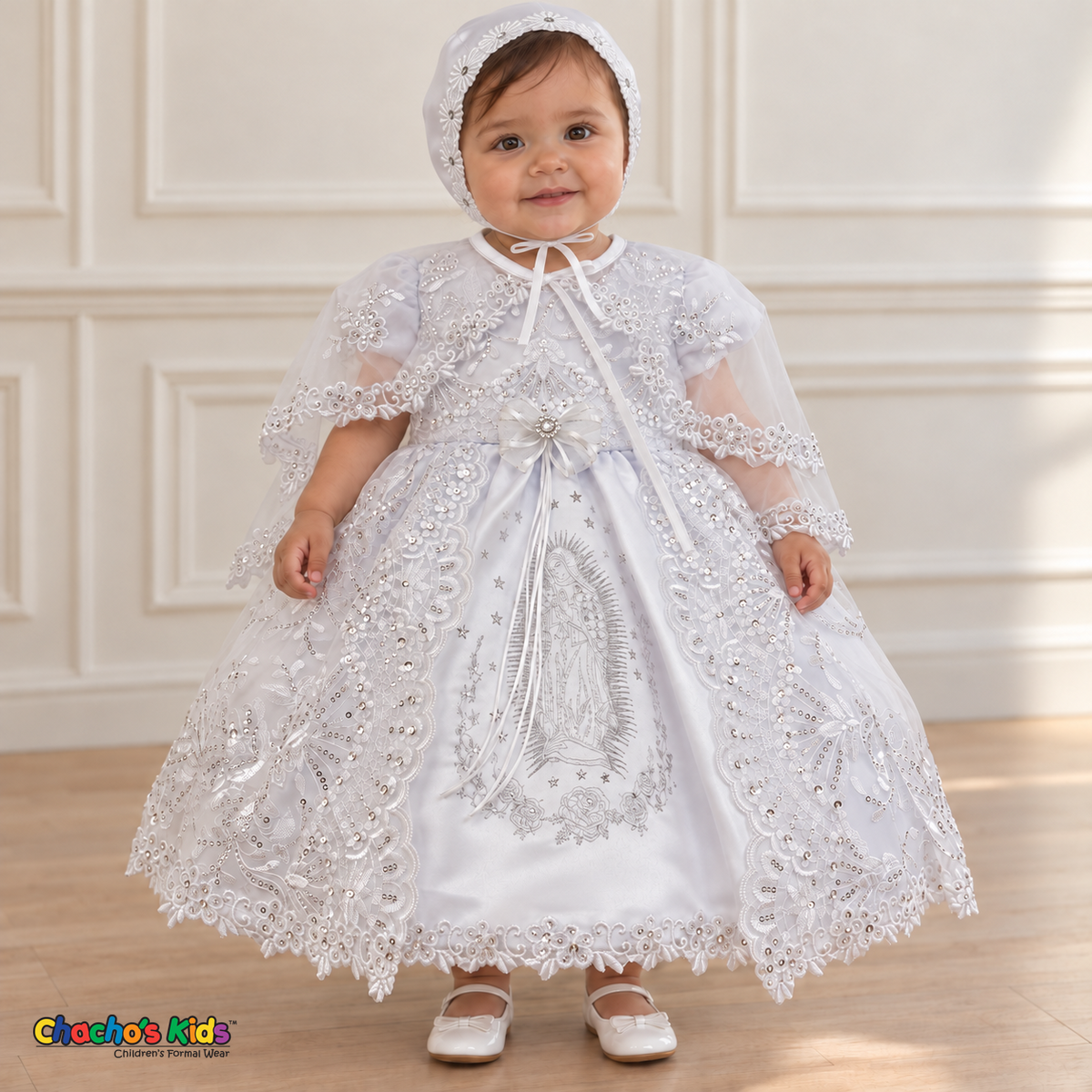 Baby girl wearing the white Virgin Mary Baptism Dress, Style 374, and matching satin bonnet, front view with the sheer embroidered cape, showing silver Virgin Mary embroidery and floral lace.