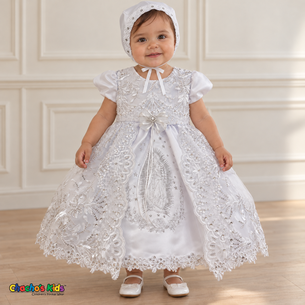 Baby girl wearing the white Virgin Mary Baptism Dress, Style 374, and matching satin bonnet, front view without the cape, showing silver Virgin Mary embroidery and floral lace.