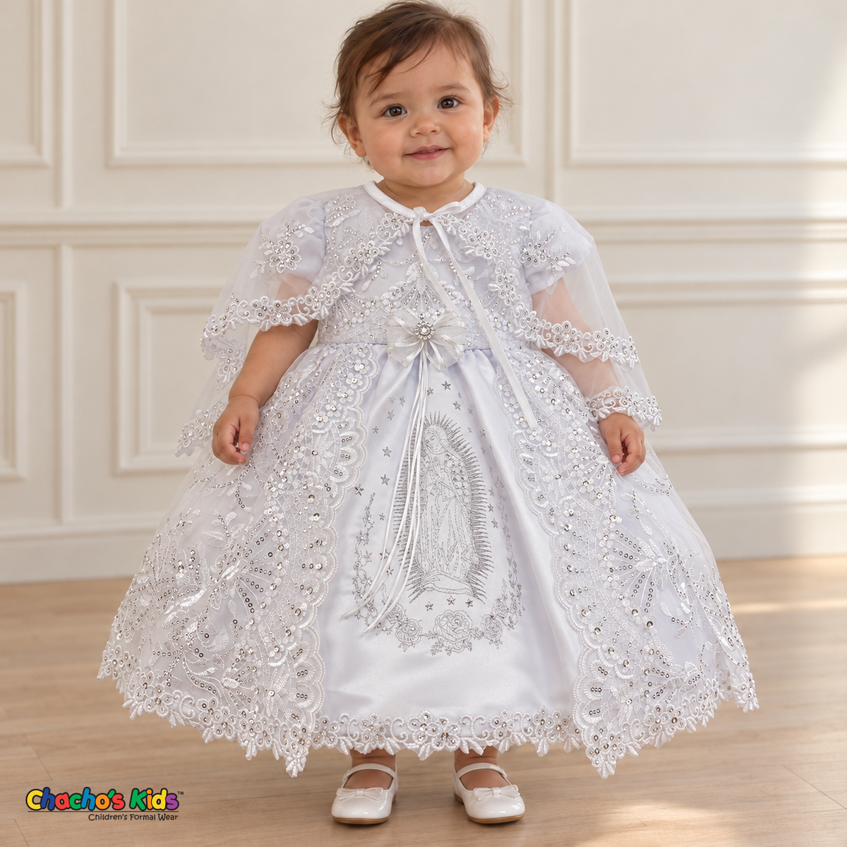 Baby girl wearing the white Virgin Mary Baptism Dress, Style 374, front with cape, showing silver Virgin Mary embroidery and floral lace.