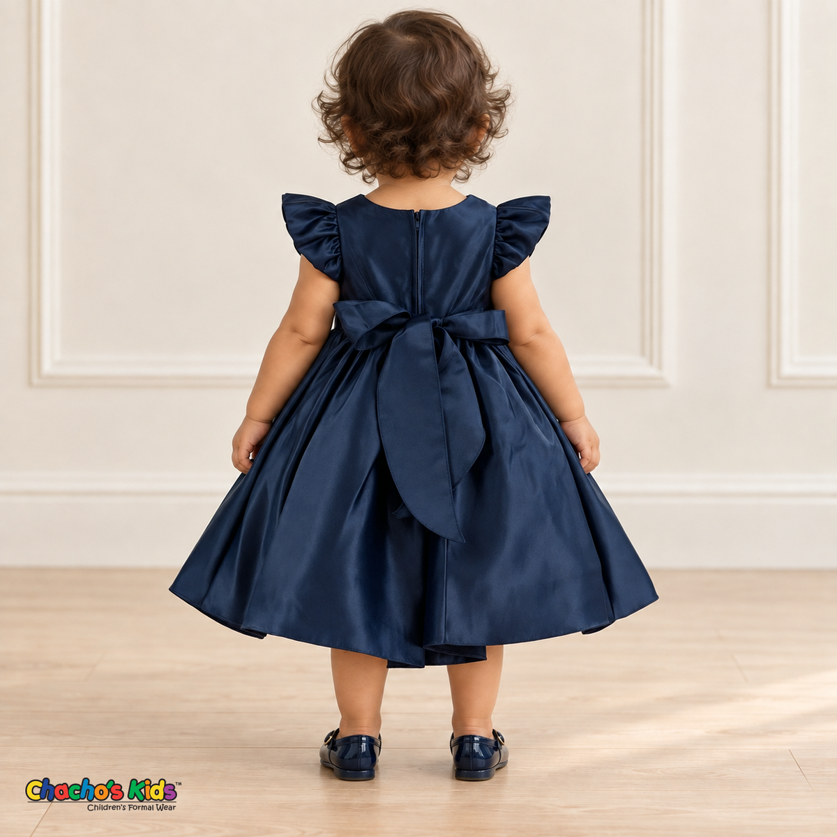 Back view of a baby girl wearing the navy blue Baby Satin Flutter & Bow Dress, Style B263, showing flutter sleeves, the back zipper and a large matching sash bow with long tails.