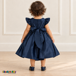 Back view of a baby girl wearing the navy blue Baby Satin Flutter & Bow Dress, Style B263, showing flutter sleeves, the back zipper and a large matching sash bow with long tails.
