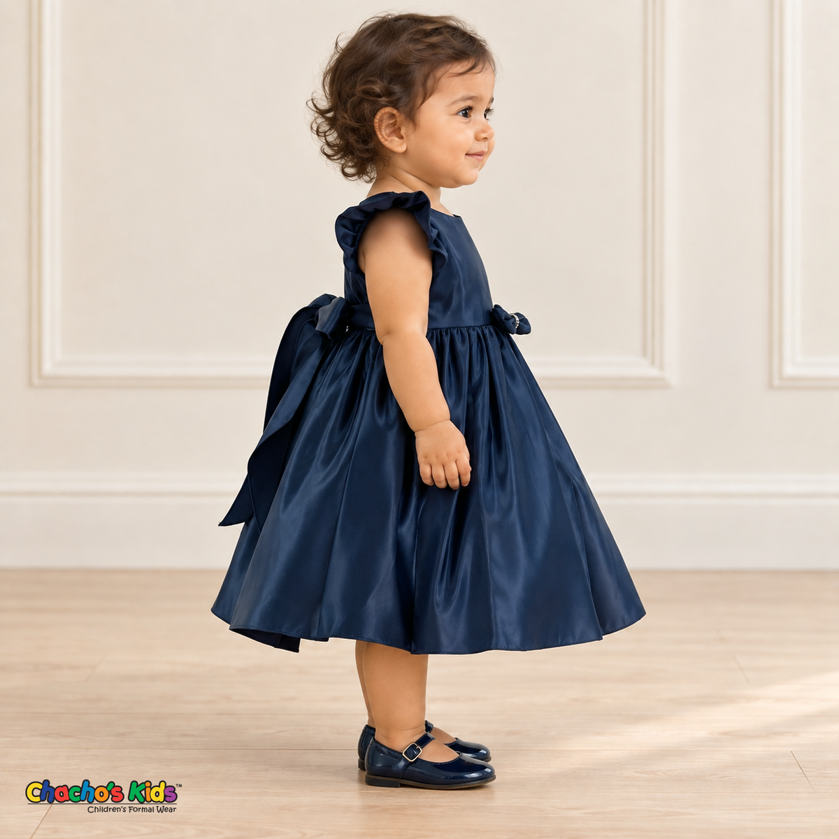 Side view of a baby girl wearing the navy blue Baby Satin Flutter & Bow Dress, Style B263, showing the flutter sleeve, gathered satin skirt, front waist bow and back sash.