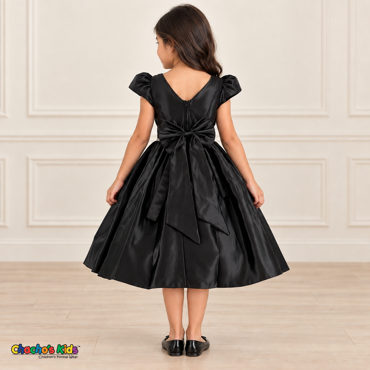 Back view of a girl wearing a black satin party dress with a V-shaped neckline, zipper and large matching satin bow.