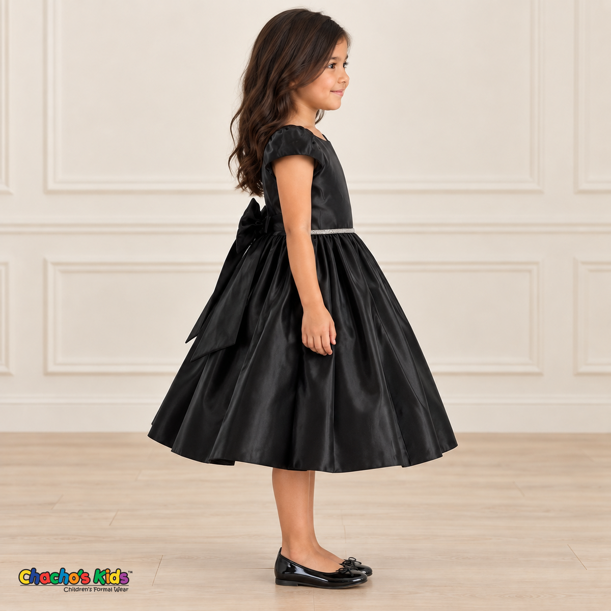 Side view of a girl wearing a black satin party dress with puff sleeves, a jeweled waist and a large back bow.