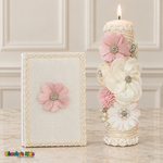Tall lit ivory baptism candle with ivory and blush-pink flowers beside an ivory Bible with a pink flower and braided edging.