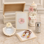 Seven-piece ivory floral baptism set with an open keepsake chest, tall lit candle, pink-flower Bible, ivory shell, rosary jar, handkerchief and visible scapular.