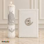 Tall lit white baptism candle with silver horse embroidery beside a white Bible with a silver horse-and-horseshoe emblem.